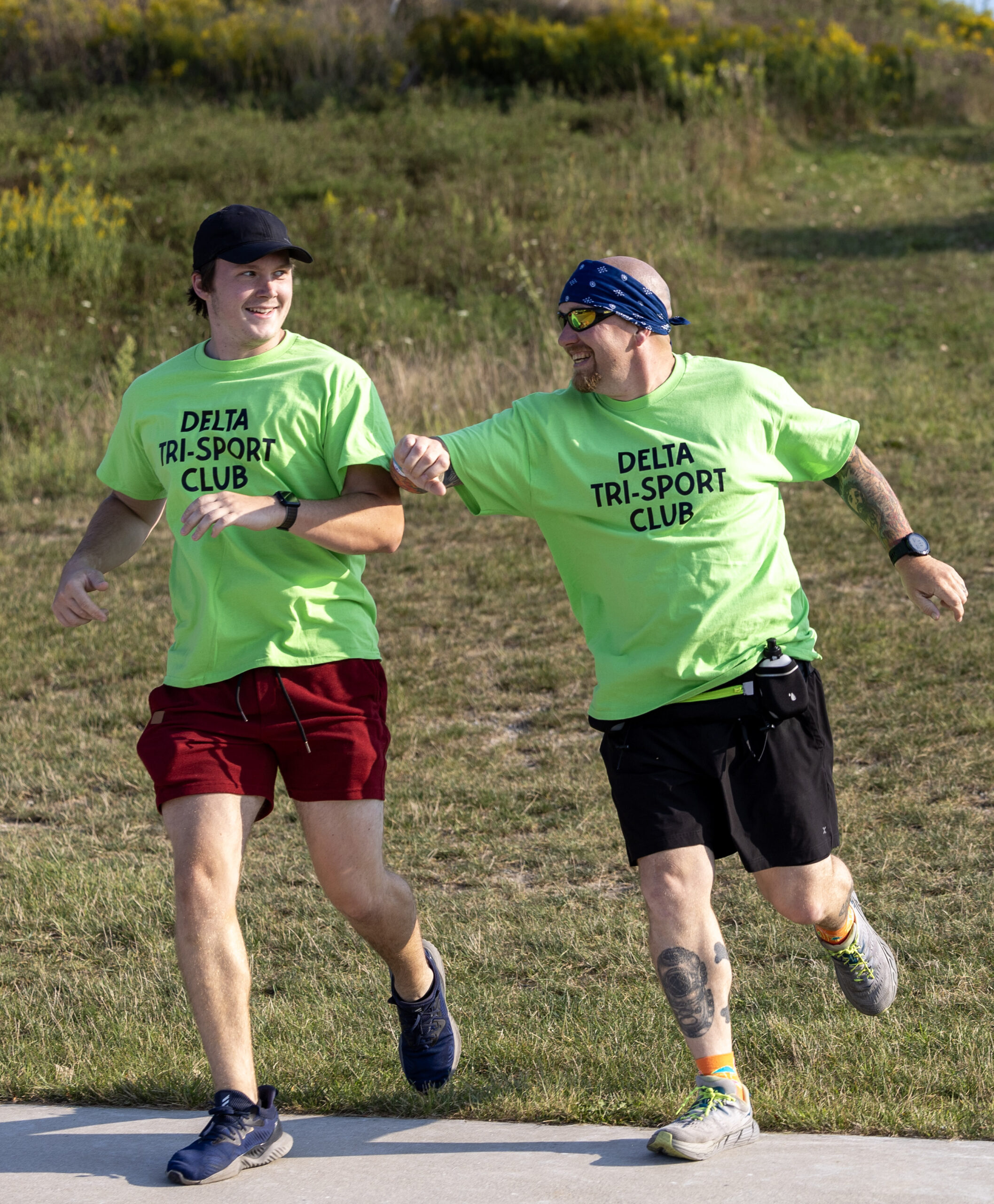 Two men wearing bright green "DELTA TRI-SPORTS CLUB" t-shirts are running and laughing together