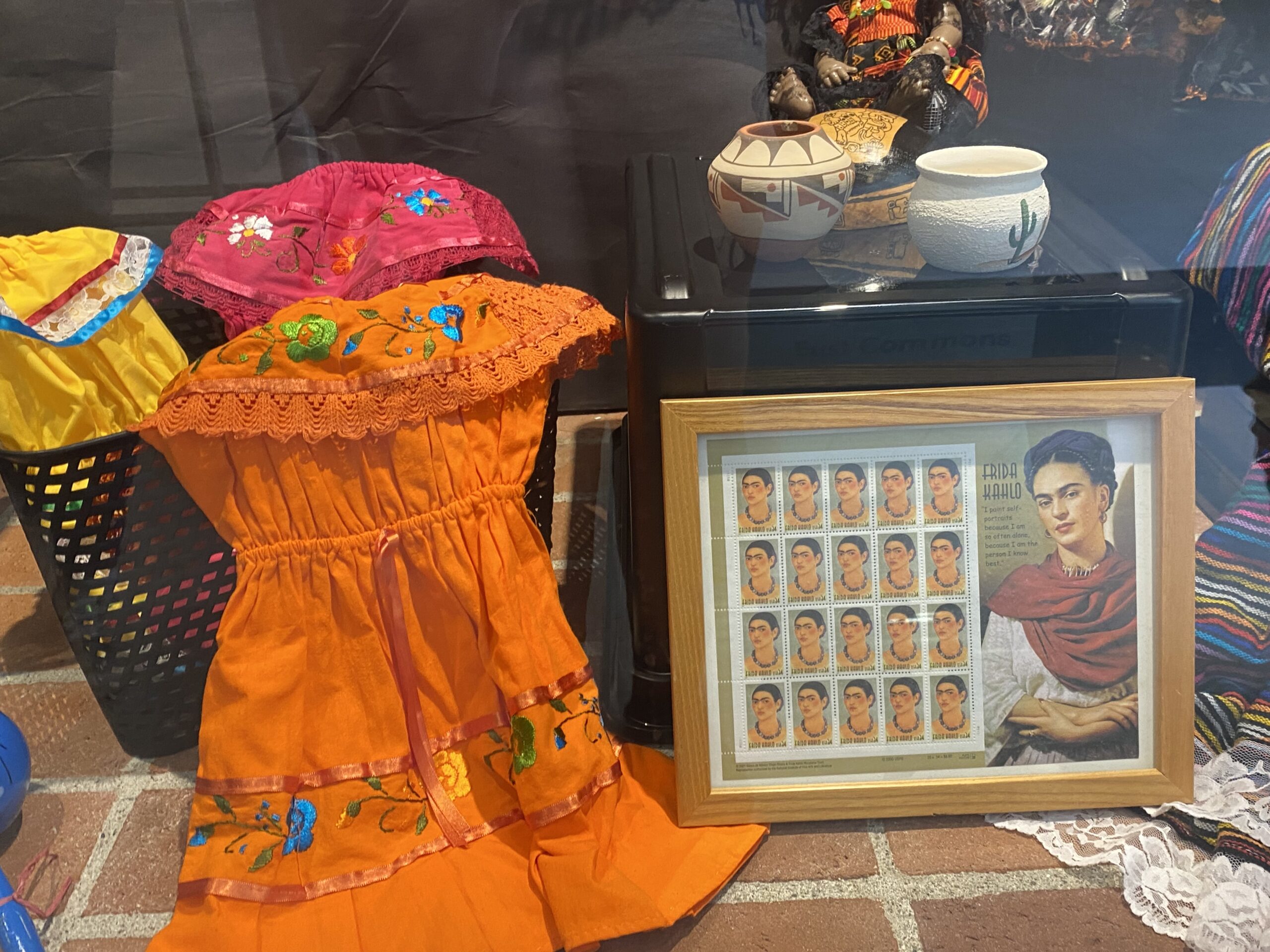 Yellow, pink and orange hispanic dresses are displayed next to a framed picture of Frida Kahlo and pottery